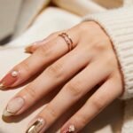 Close-up of a hand on cream satin with almond-shaped nude and ivory nails featuring gold French tips, pearl accents, and a date detail reading 07.21.1994