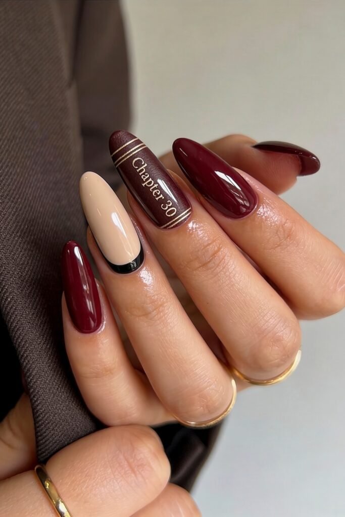 Close-up of long almond nails in glossy burgundy and nude beige, featuring one beige nail with a black French tip and one accent nail styled like a miniature book cover