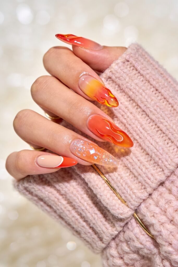 Summer Birthday Nails That Feel Fresh and Fun 1 Hand in a pink knit sleeve showing long orange and yellow flame nails with one glitter accent nail and a bright orange French tip