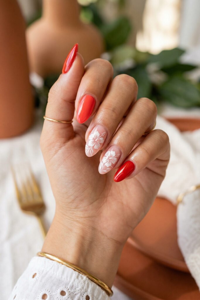 Almond nails with white hibiscus designs on sheer nude polish and glossy coral red accents styled in a warm sunlit indoor setting