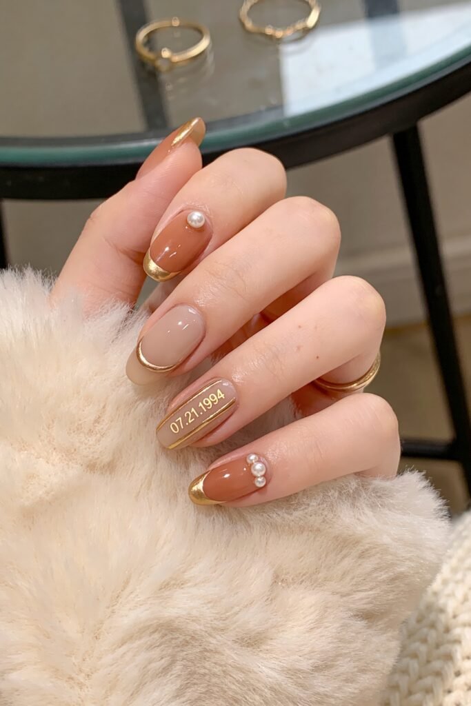 Gold Birthday Nails That Feel So Chic 20 Close-up of almond-shaped nails in caramel brown and taupe with gold French tips, pearl accents, and a date detail reading 07.21.1994