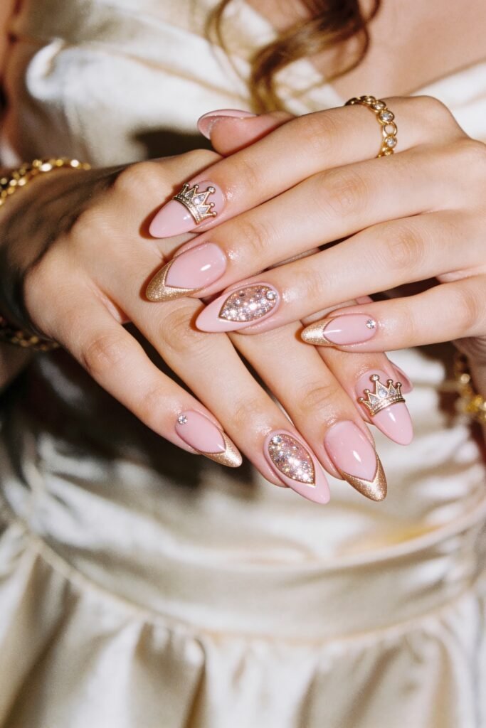 Two hands with glossy blush pink almond nails featuring gold glitter French tips, rhinestone crown charms, and crystal-encrusted accent nails