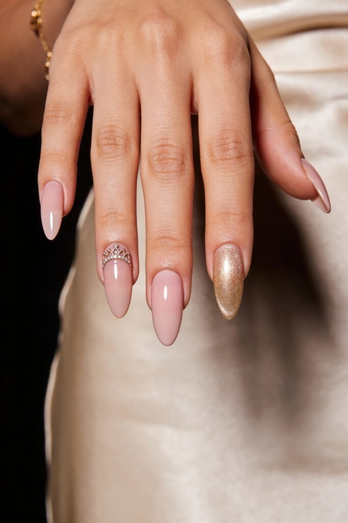 Close-up of a hand with glossy pale pink almond nails, featuring a crown charm accent and one full gold glitter nail