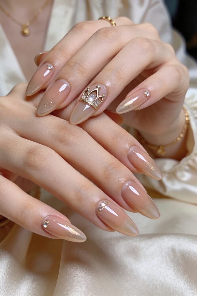 Two hands with glossy nude beige ombré almond nails, accented with small rhinestones and a jeweled crown charm