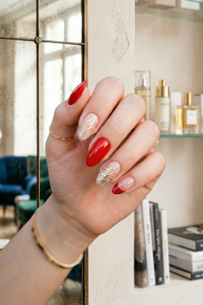 Close up of glossy red almond nails with nude accent nails featuring gold glitter details and a red French tip against a mirrored shelf backdrop