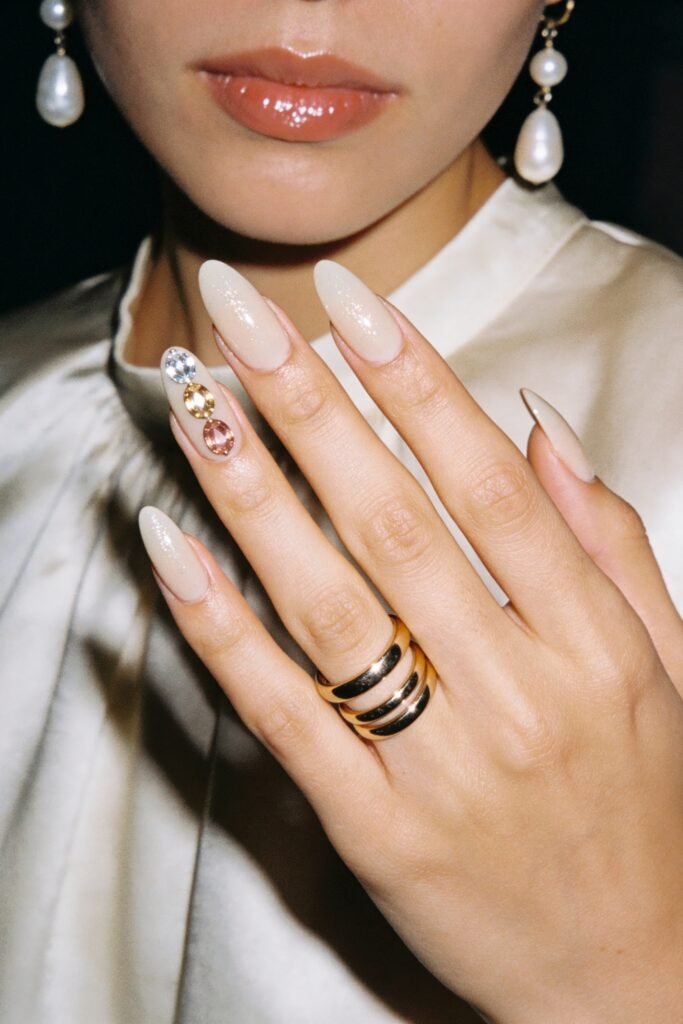 Close-up of milky white almond nails with a glossy shimmer and one accent nail decorated with three oval gemstones
