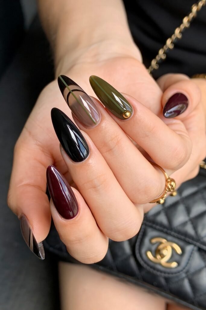 Close-up of long almond nails in glossy black, burgundy, olive green, and smoky taupe, with one striped accent nail and a small gold stud detail beside a black quilted handbag