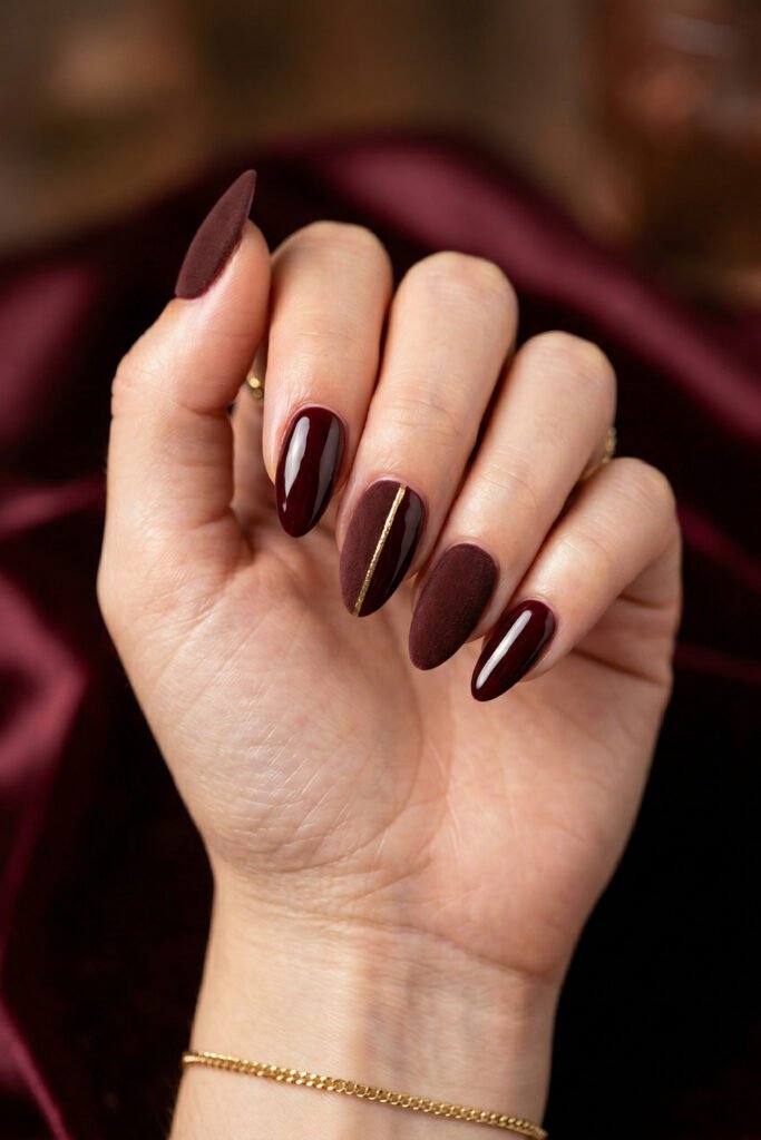 Brown And Red Nails That Make Every Look Richer 3 Hand with glossy burgundy and matte plum almond nails featuring a thin gold stripe accent against a rich satin background