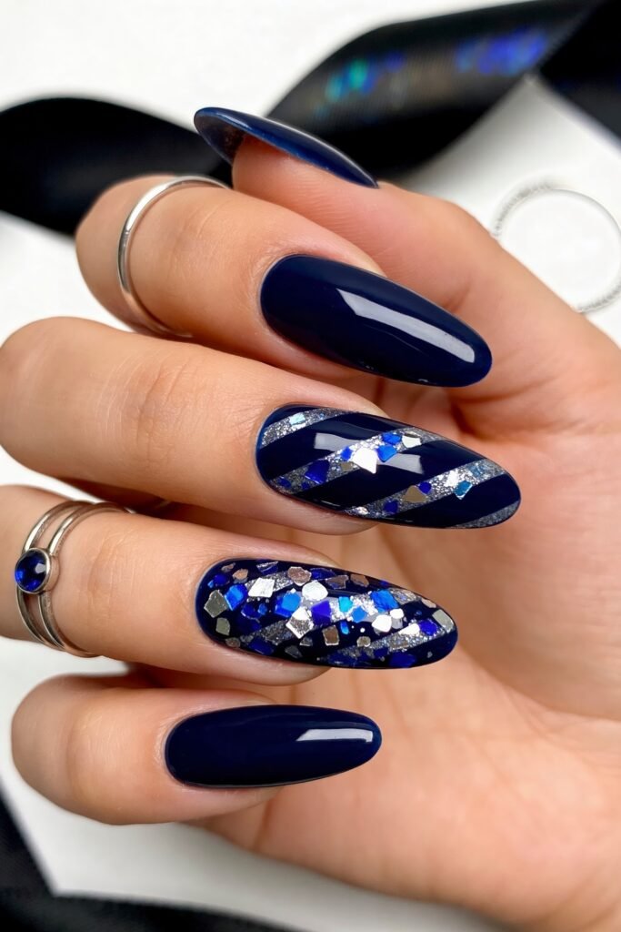 Close-up of glossy dark navy nails with silver and blue glitter accents, including one striped nail and one densely packed confetti glitter nail