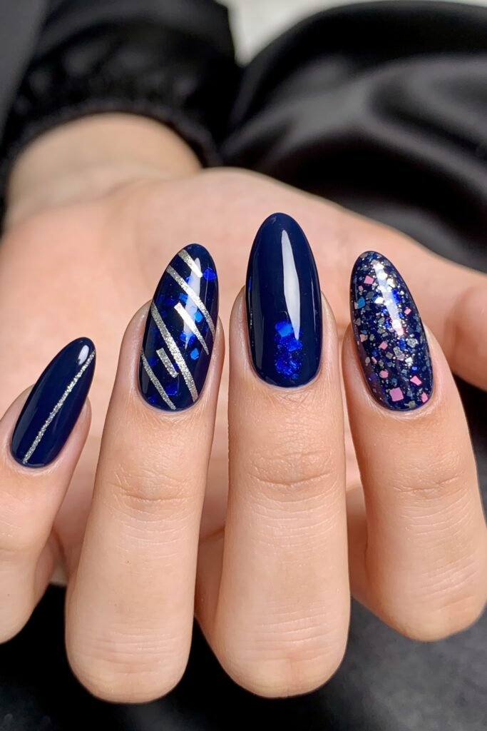 Hand with glossy deep navy nails featuring silver diagonal lines, blue foil accents, and one full glitter accent nail