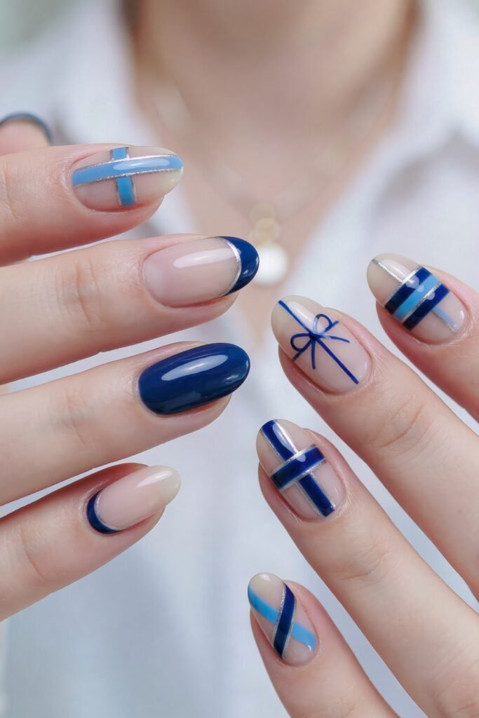 Close-up of short nude nails with navy and light blue geometric line art, silver accents, and one nail decorated like a tied ribbon bow
