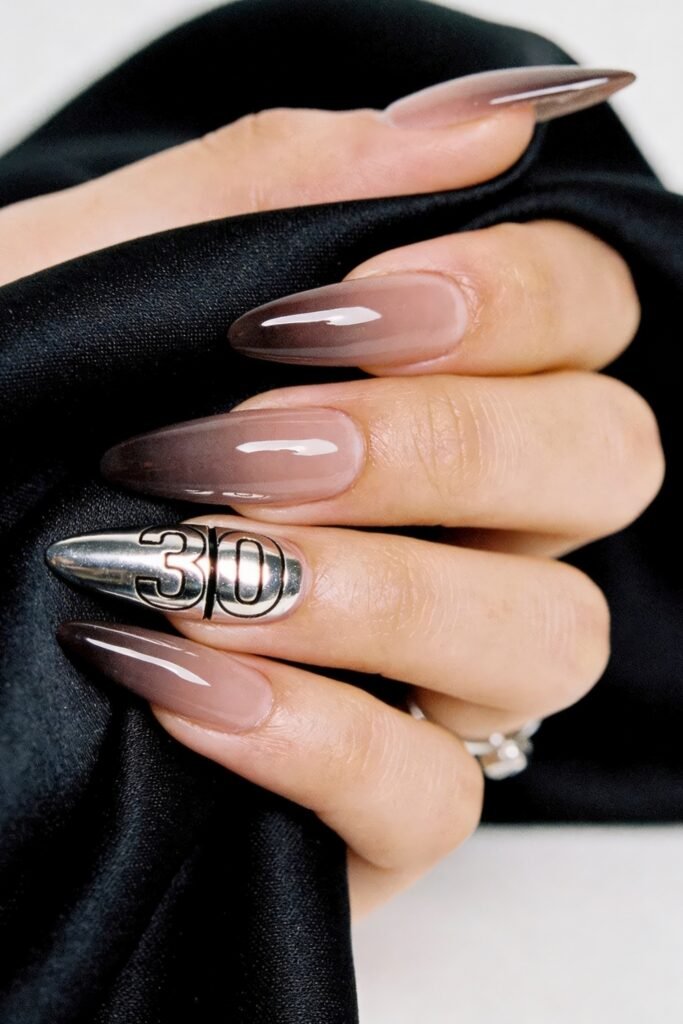 Close-up of glossy taupe ombré almond nails with one silver chrome accent nail, holding black satin fabric