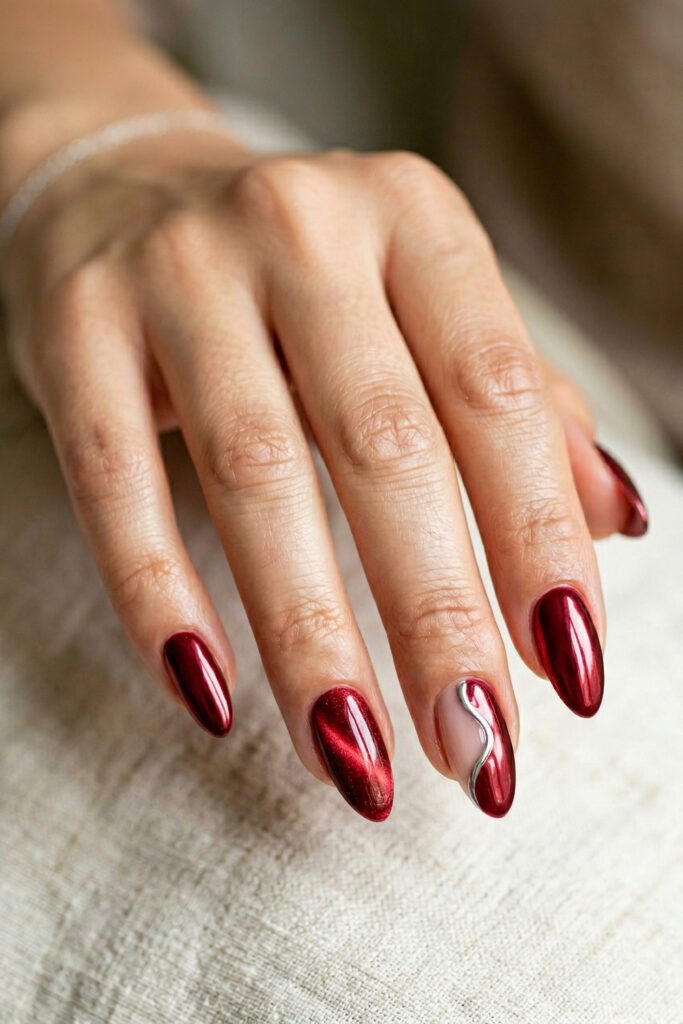 Deep red metallic almond nails with a silver wave accent on one nude nail styled in a soft indoor close up