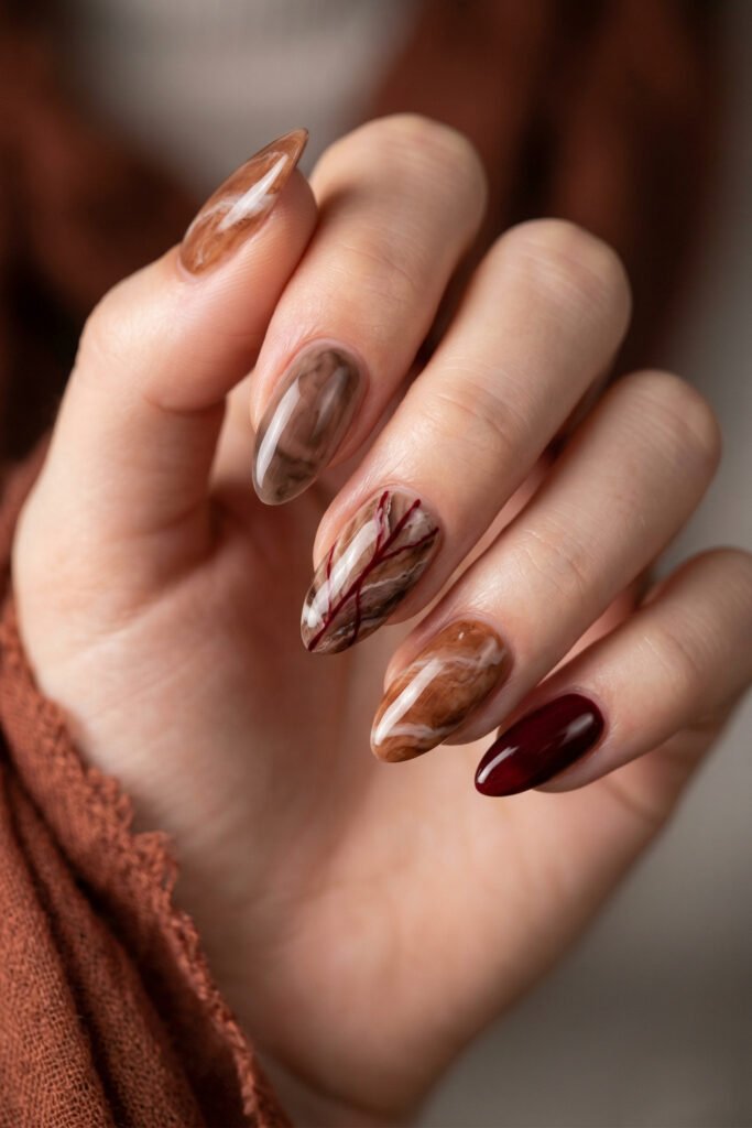 Brown And Red Nails That Make Every Look Richer 9 Close up of almond nails with smoky taupe and caramel marble designs plus a glossy burgundy accent nail against a warm blurred background