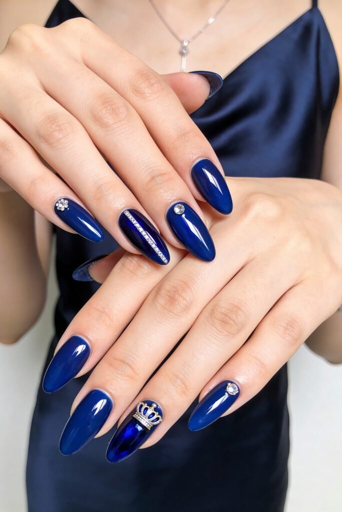 Both hands with glossy royal blue almond nails featuring rhinestones, a silver stripe accent, and a jeweled crown charm on one nail