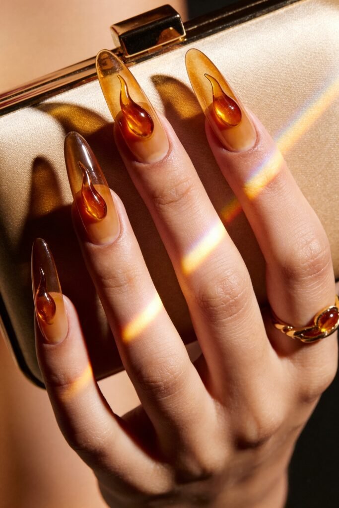 Close-up of long translucent amber almond nails with glossy raised flame accents, posed against a metallic clutch with rainbow light reflections