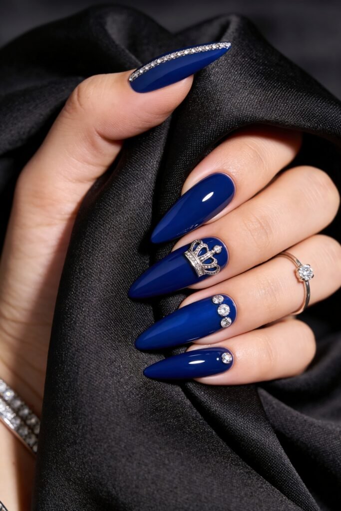 Close-up of long royal blue stiletto nails with crystal accents, a silver crown embellishment, and a rhinestone-lined thumb against black satin