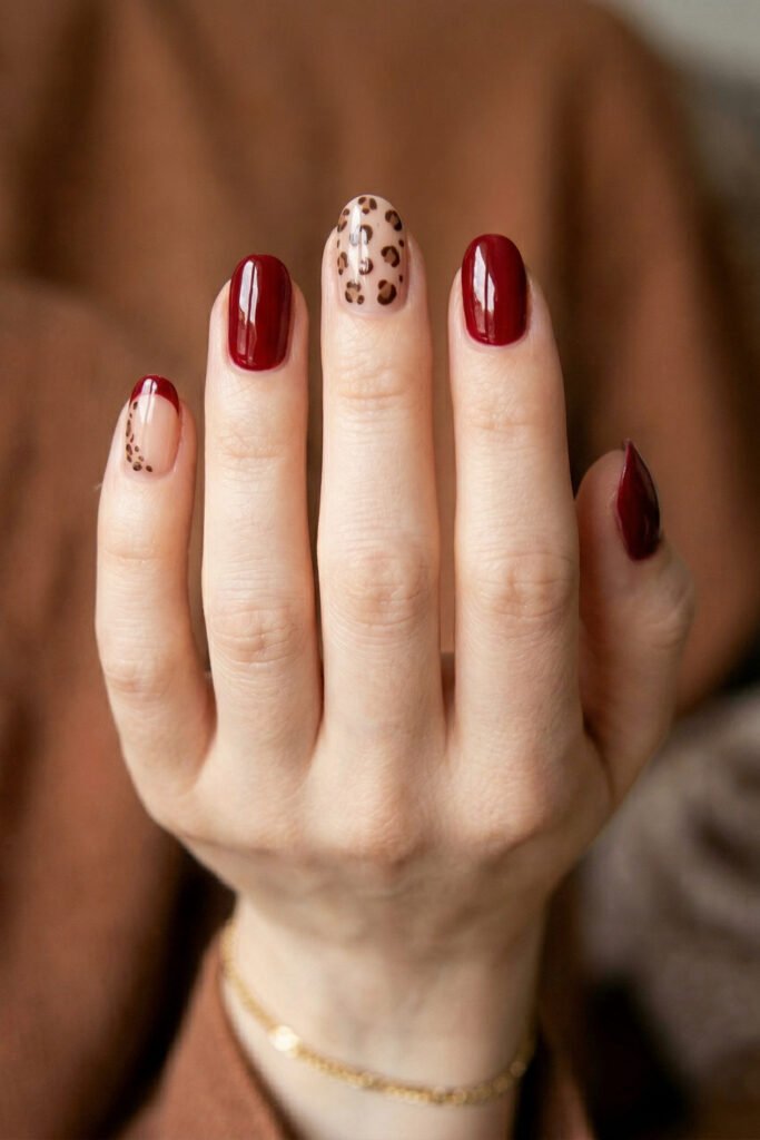 Brown And Red Nails That Make Every Look Richer 11 Hand with glossy burgundy nails featuring a nude leopard accent nail and a leopard French tip against a warm blurred background