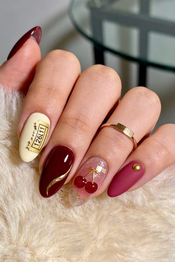 Gold Birthday Nails That Feel So Chic 13 Close-up of almond nails in ivory, burgundy, and berry tones with a ticket detail reading 126L11, a gold swoosh, translucent pink glitter, and raised red cherry accents