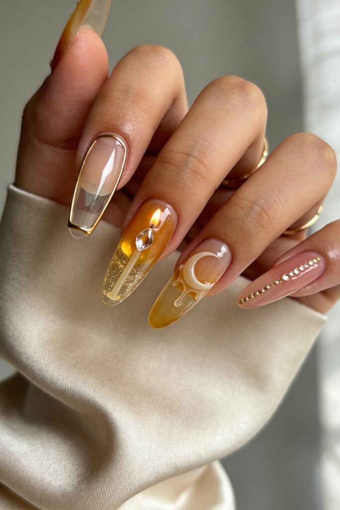 Gold Birthday Nails That Feel So Chic 16 Close-up of glossy almond nails in nude, amber, and blush tones featuring a clear gold-outlined tip, a lit candle design with glitter, a white crescent with dripping detail, and a rhinestone stripe