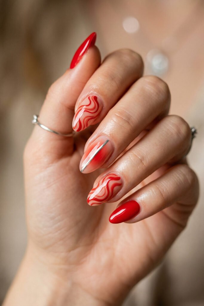 Almond nails with glossy red polish abstract red swirl art on nude bases and a silver stripe accent in warm light