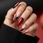 Chic Gold and Red Nails for a Bold, Polished Look Close up of a hand with gold rings and glossy almond nails in red black and nude featuring sharp French tips and a tiny heart accent