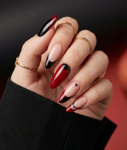 Close up of a hand with gold rings and glossy almond nails in red black and nude featuring sharp French tips and a tiny heart accent