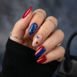 Glossy almond blue and red nails with raised star art, a sheer nude accent nail with red and blue stars, and silver rings on hand