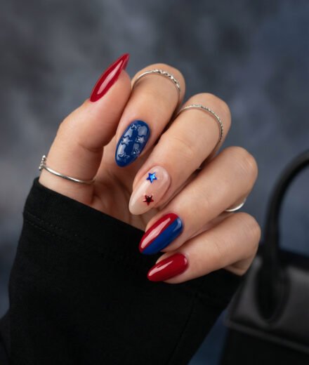 Glossy almond blue and red nails with raised star art, a sheer nude accent nail with red and blue stars, and silver rings on hand