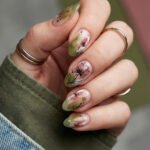 Olive green and sheer almond edgy spring nails with pink floral art, black speckles, and delicate leaf details on a natural nude base.
