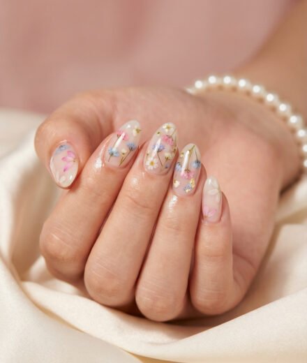 Glossy almond floral spring nails with sheer milky nude base, pressed pink, blue, and white flowers, and subtle gold foil on a soft satin background.