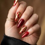 Brown And Red Nails That Make Every Look Richer Close up of a hand with glossy red almond nails and gold foil accent nails with delicate rings and a black satin sleeve against a warm wood background