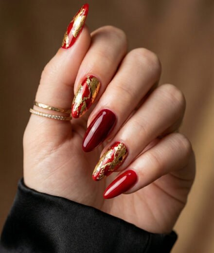 Close up of a hand with glossy red almond nails and gold foil accent nails with delicate rings and a black satin sleeve against a warm wood background
