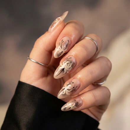 Spring Goth Nails That Make Dark Romance Feel Fresh Almond nude nails with white floral art and fine black accents, a soft dark-romance take on spring goth nails with a glossy neutral base and elegant stiletto shape.
