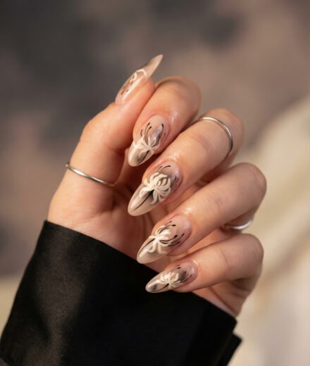 Almond nude nails with white floral art and fine black accents, a soft dark-romance take on spring goth nails with a glossy neutral base and elegant stiletto shape.