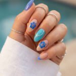 Blue almond spring summer nails with hibiscus flower art, turquoise accent nail, silver wave detail, and blue French tip against a poolside background.