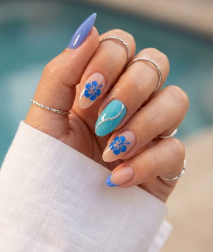 Blue almond spring summer nails with hibiscus flower art, turquoise accent nail, silver wave detail, and blue French tip against a poolside background.