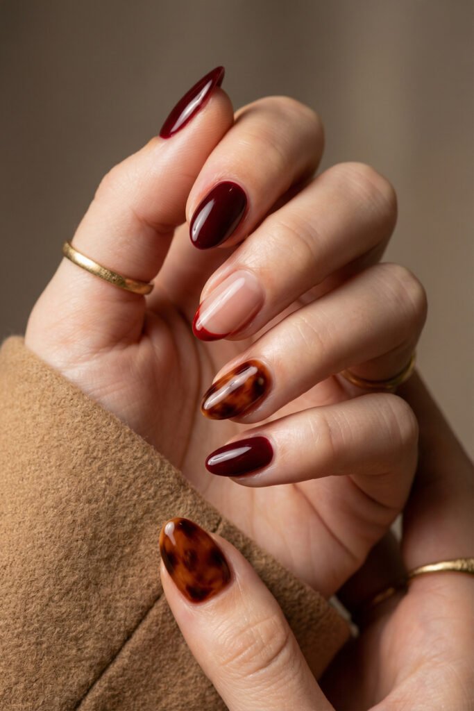 Almond fall red nails with glossy burgundy polish, a nude red French tip, and tortoiseshell accent nails for a rich autumn manicure.
