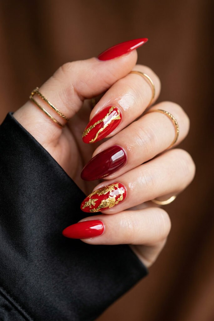 Chic Gold and Red Nails for a Bold, Polished Look 1 Close up of a hand with glossy red almond nails featuring gold foil accents and a deep burgundy nail with slim gold rings and a black satin sleeve