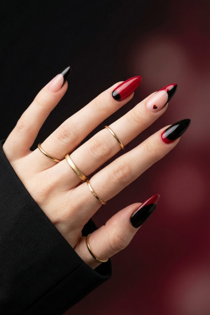 Black and Red Nails for a Bold, Polished Look 2 Hand with gold rings displaying red black and nude almond nails with sleek color blocks and a small heart accent against a dark blurred background