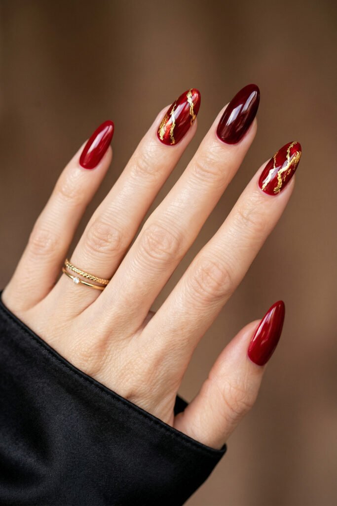 Chic Gold and Red Nails for a Bold, Polished Look 2 Hand with glossy red and burgundy almond nails featuring gold foil accent designs with stacked rings and a black sleeve against a warm wood background
