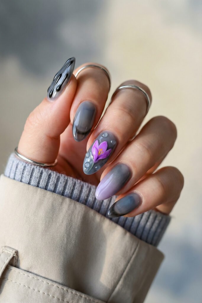 Moody gray edgy spring nails with black drip accents, soft lilac ombré, clear droplet details, and a purple crocus flower on glossy almond nails.
