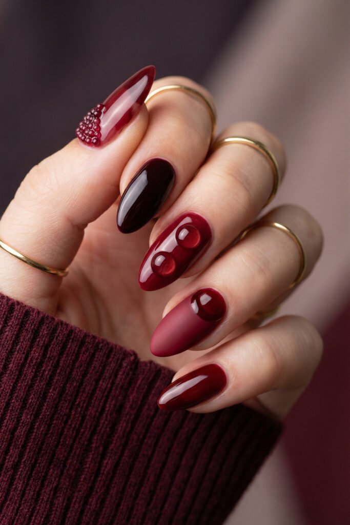 Glossy fall red nails with deep burgundy tones, 3D bubble accents, a matte berry gradient nail, and a beaded detail on the thumb.
