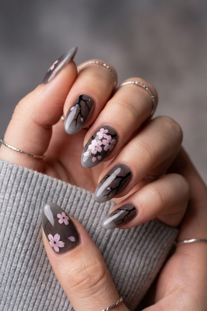 Smoky gray almond nails with black branch art and pale pink cherry blossoms, creating an elegant spring goth nails look with a misty floral finish.
