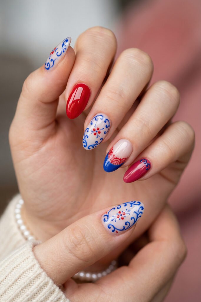 Elegant blue and red nails with ornate blue scroll art, tiny floral accents, glossy red polish, and a refined almond shape
