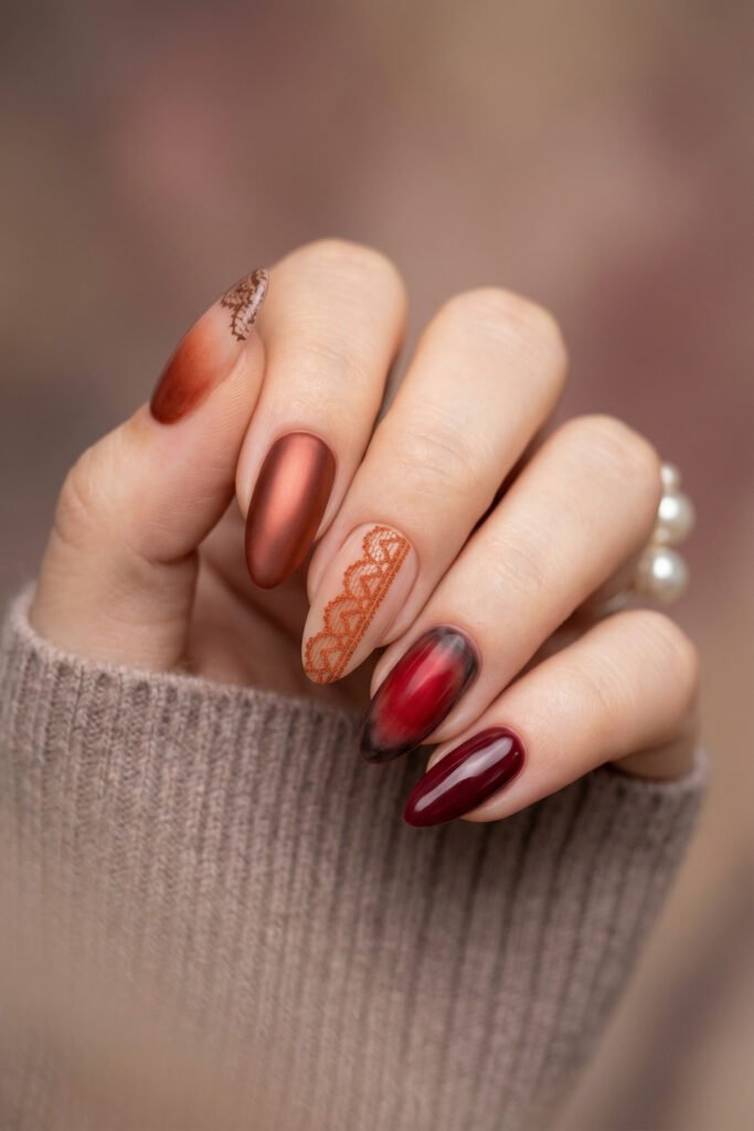 Almond fall red nails with matte copper, lace-inspired nude art, a smoky red aura accent, and glossy burgundy polish.
