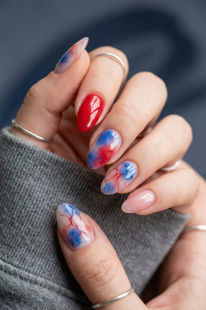 Almond blue and red nails with soft watercolor marble details, fine veining, one glossy red accent nail, and a sheer pink nail
