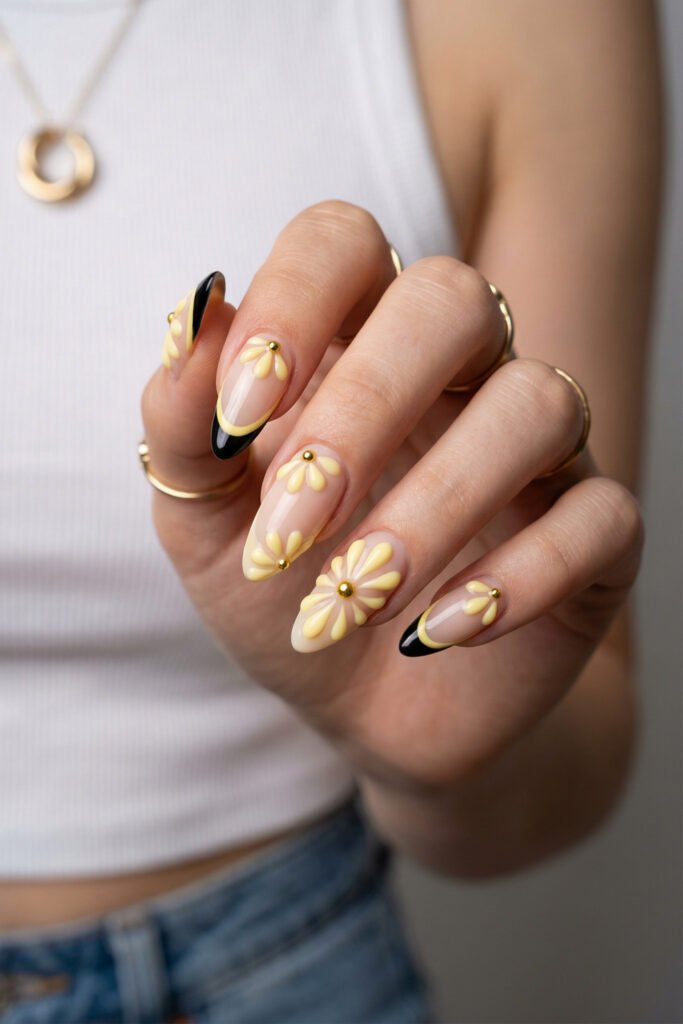 Yellow daisy edgy spring nails with 3D floral details, gold centers, and black French tips on long almond nude nails.
