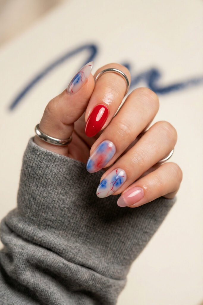 Fresh almond blue and red nails featuring cloudy marble art, a glossy red accent, sheer pink nude nail, and cozy gray sleeve
