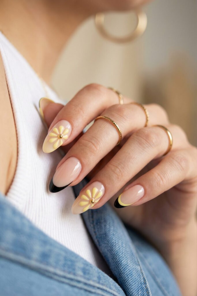 Soft nude edgy spring nails with yellow daisy accents and black French tips on glossy almond nails for a playful spring manicure.
